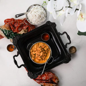 bowl of indian butter chicken on a black tray with white rice on the side placed on top of a decorative shawl flat lay