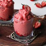 Two glasses filled with scoops of strawberry nice cream topped with fresh strawberries and displayed on a wooden board