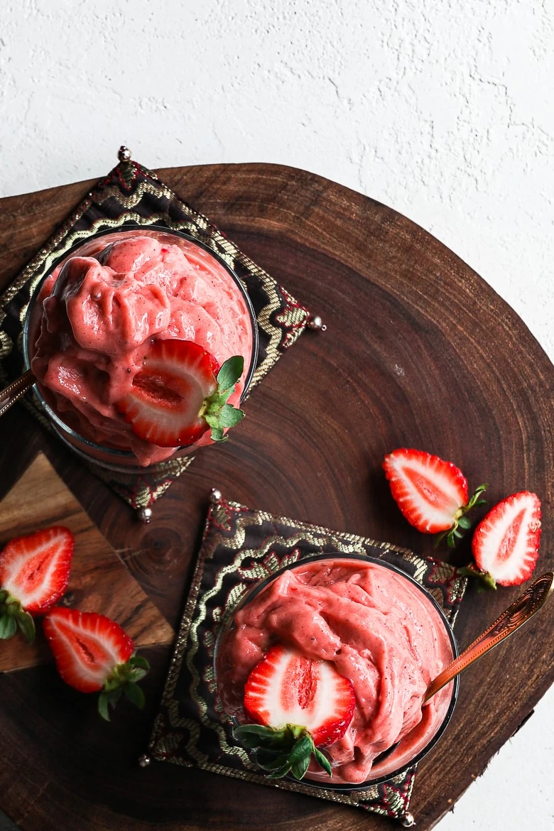 flat-lay: two glasses filled with scoops of strawberry nice cream topped with fresh strawberries and displayed on a wooden board