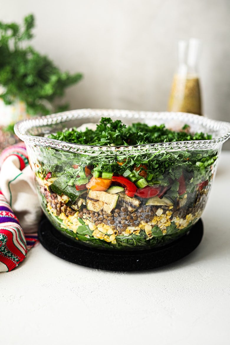 glass bowl with layers of grilled vegetables and leafy greens with a dressing bottle in the background