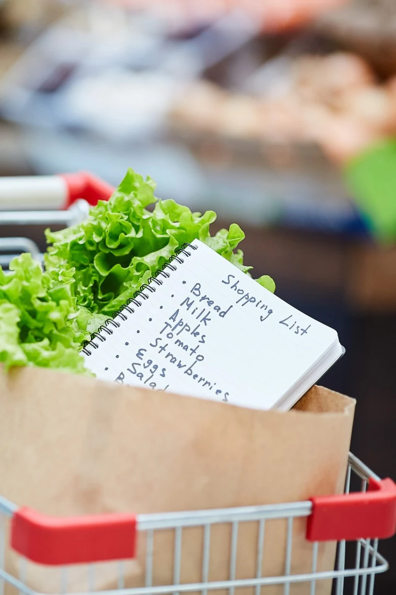 Background image of shopping cart with fresh groceries, focus on shopping list in paper bag, copy space