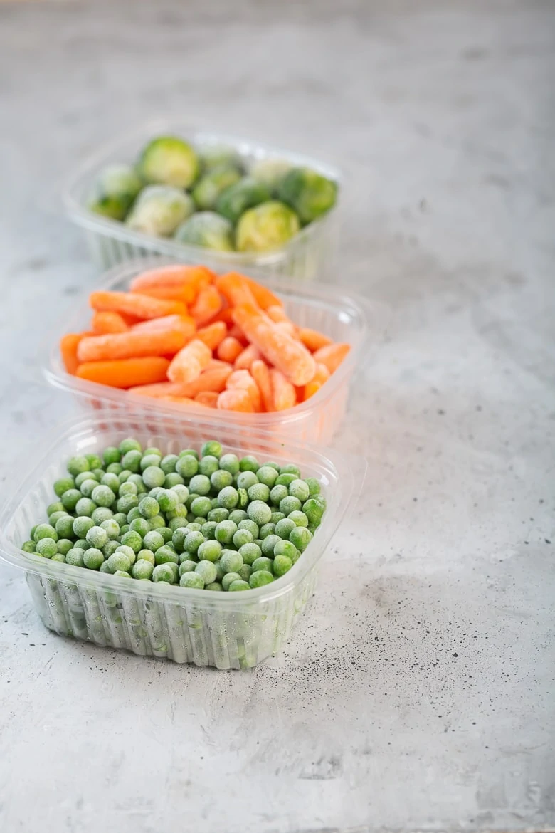 Frozen vegetables such as green peas, brussels sprouts and baby carrot in the storage boxes on the concrete gray background, close up