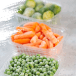 Frozen vegetables such as green peas, brussels sprouts and baby carrot in the storage boxes on the concrete gray background