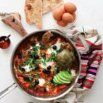 a pan of shakshuka with eggs garnished with olives, herbs, avocado slices and artichoke, with a bowl of raw eggs and naan bread close by - flatlay