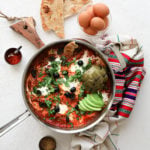 close up of a pan of shakshuka with eggs garnished with olives, herbs, avocado slices and artichoke, with a bowl of raw eggs and naan bread close by - flatlay