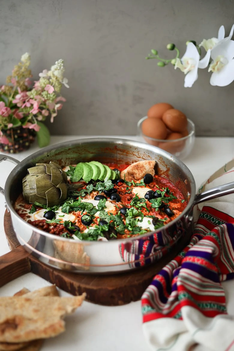 a pan of shakshuka with eggs on a wooden board garnished with olives, herbs, avocado slices and artichoke, with a bowl of raw eggs and flowers in the background - perspective