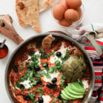 close up of a pan of shakshuka with eggs garnished with olives, herbs, avocado slices and artichoke, with a bowl of raw eggs and naan bread close by - flatlay