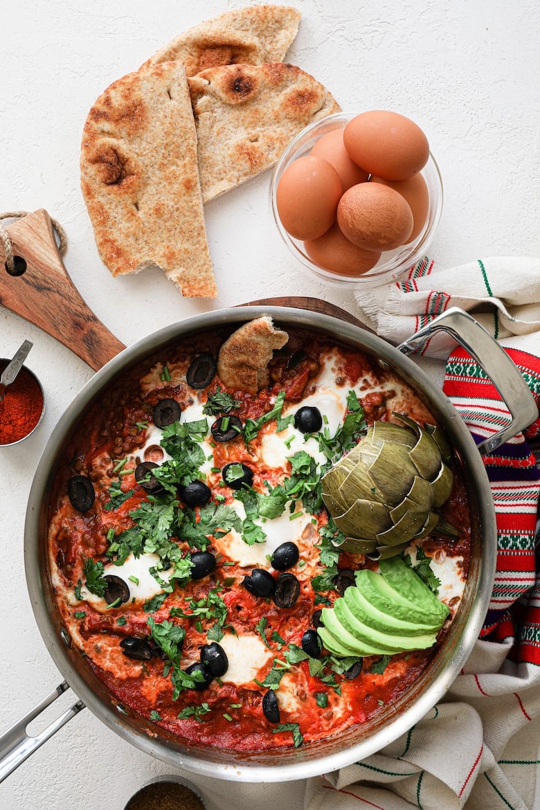 close up of a pan of shakshuka with eggs garnished with olives, herbs, avocado slices and artichoke, with a bowl of raw eggs and naan bread close by - flatlay