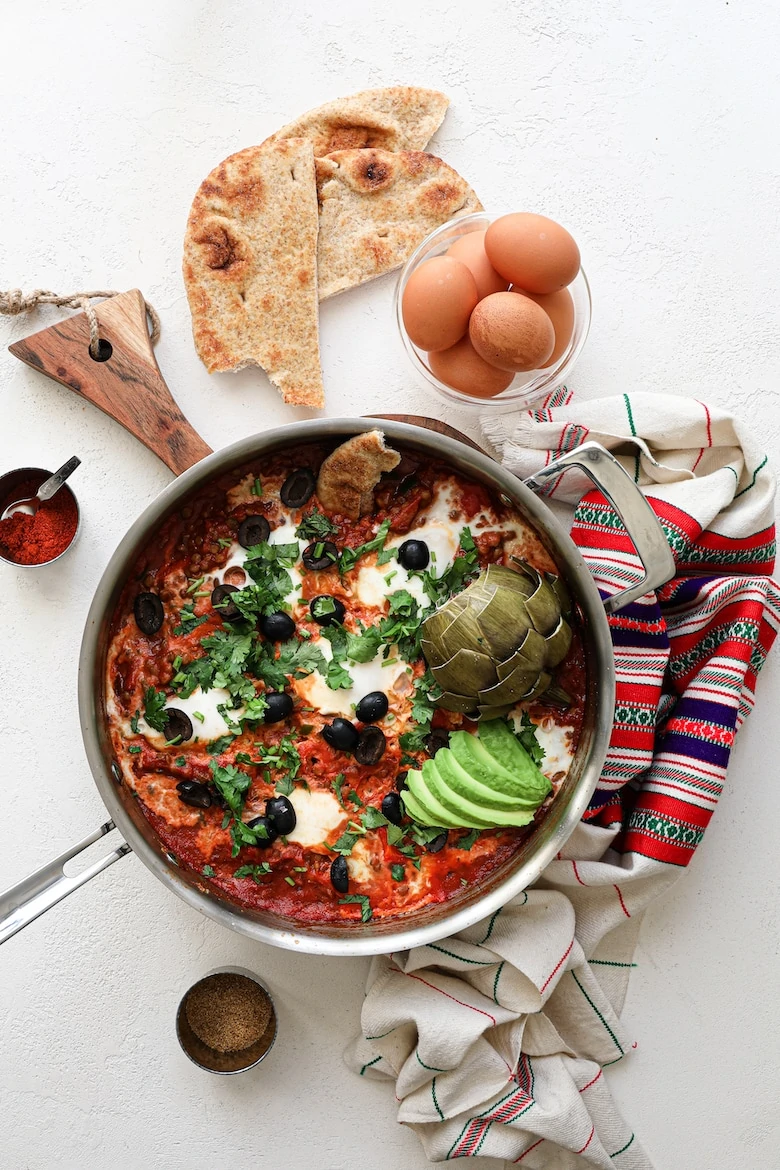 a pan of shakshuka with eggs garnished with olives, herbs, avocado slices and artichoke, with a bowl of raw eggs and naan bread close by - flatlay