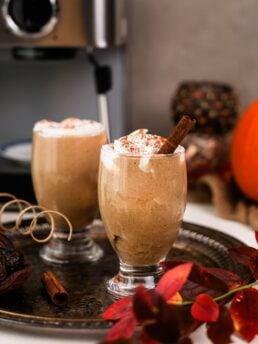 two glasses of iced pumpkin spice latte on a tray with an espresso machine in the background and other fall props