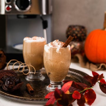 two glasses of iced pumpkin spice latte on a tray with an espresso machine in the background and other fall props