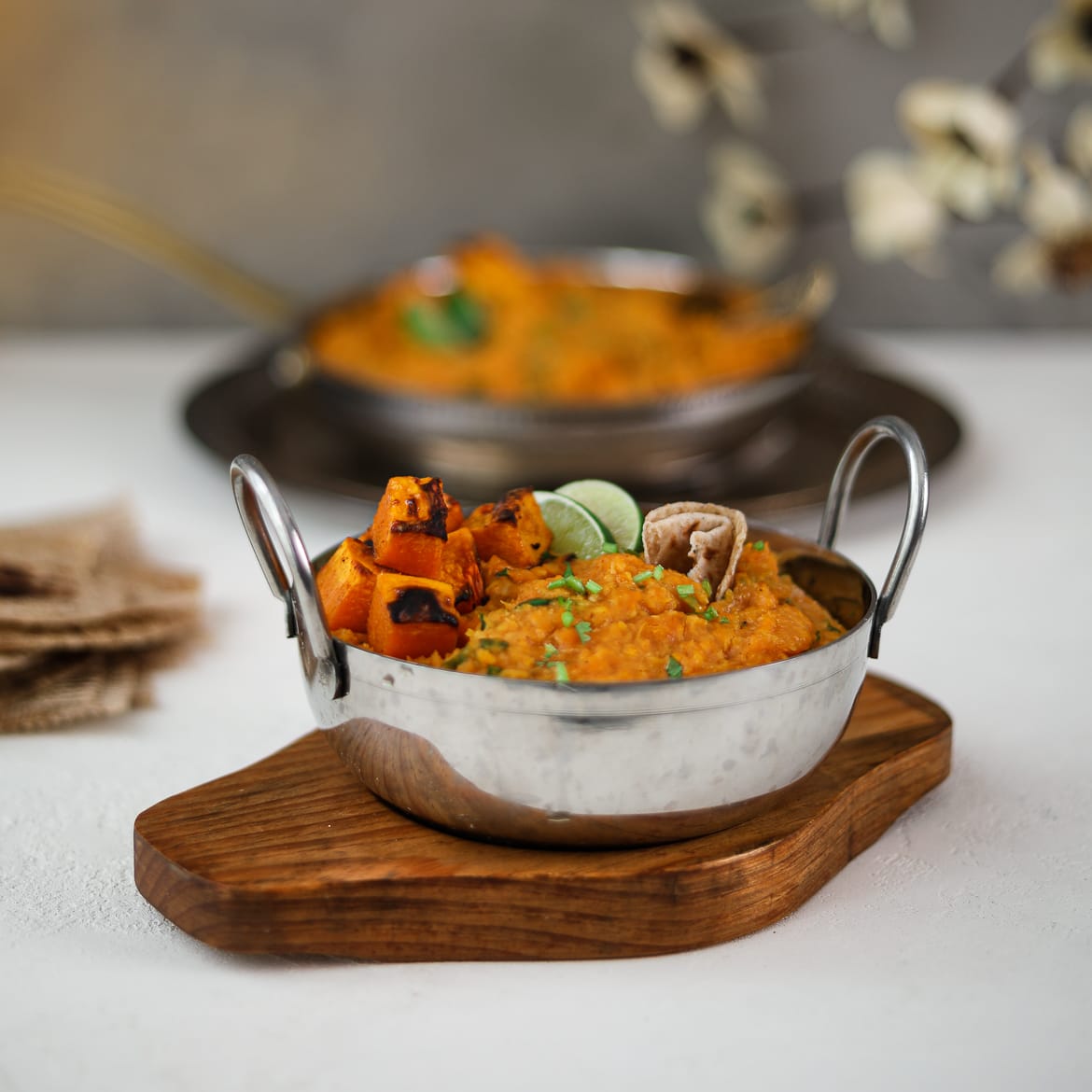 Two bowls of yellow daal topped with squash and limes wedges placed on trays