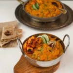 Two bowls of yellow daal topped with squash and limes wedges placed on trays with roti on the side