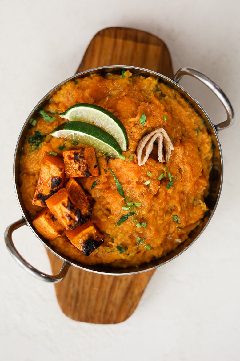 a bowl of yellow daal topped with butternut squasg and lime wedges placed on a wooden tray