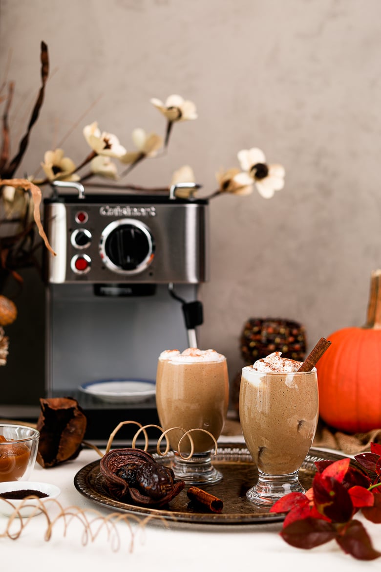 two glasses of iced pumpkin spice latte on a tray with an espresso machine in the background and other fall props