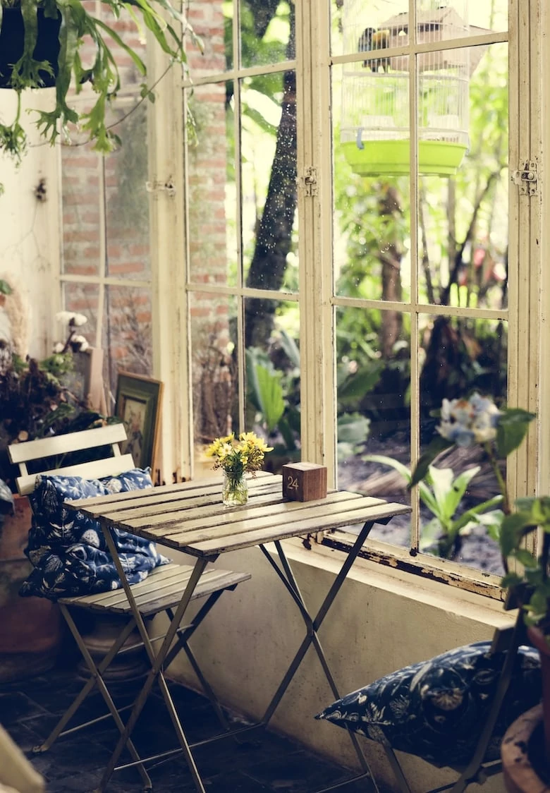 A relaxed cafe scene with wooden table and 2 chairs near a window overlooking a garden space