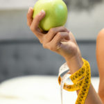 Woman holding a fresh healthy green apple in her hand with a tape measure wound round her arm in a diet and weight loss concept in close up
