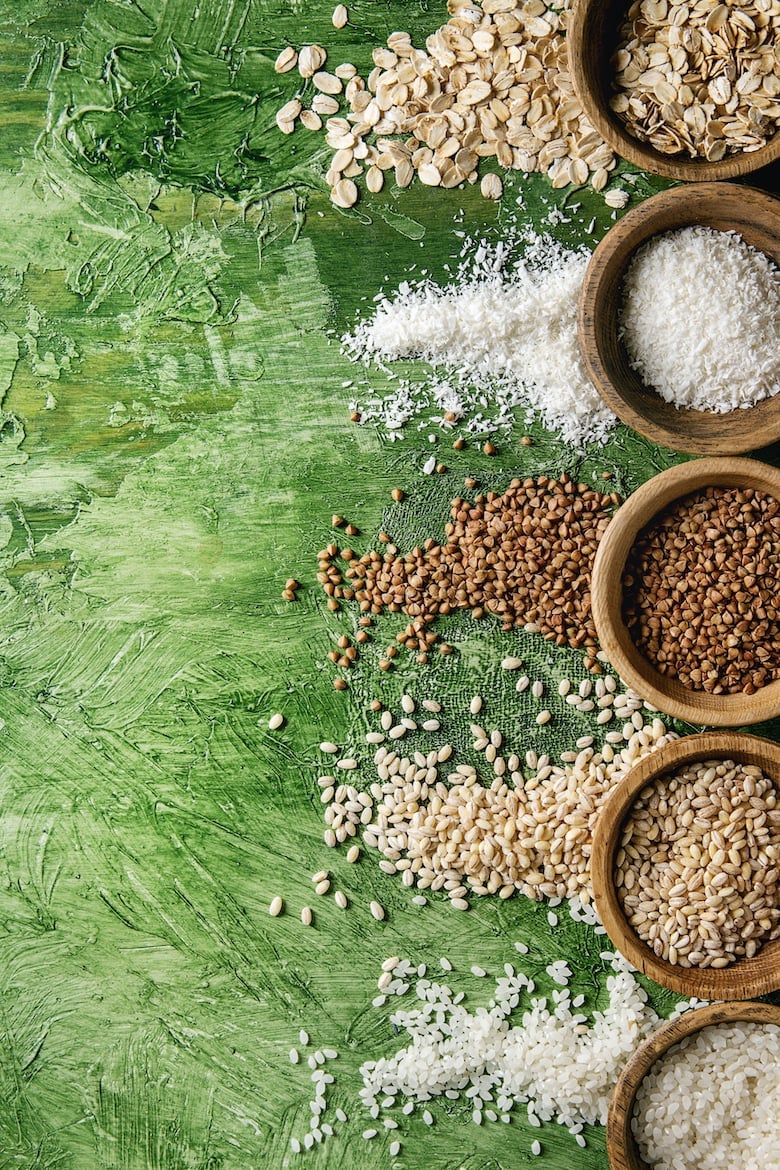 Variety of raw uncooked grains superfood cereal linen seeds, sesame, mung bean, wheat, buckwheat, oatmeal, coconut, rice in wooden bowls over green texture background. Flat lay, space