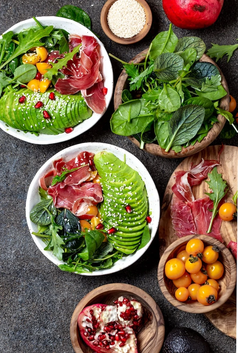 Low carbs bowl. Fresh salad with green spinach, rucola, avocado an ham serrano in white bowl, gray background, top view.