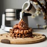 pouring maple syrup on a stack of brown waffles topped with baked pears and pecans with a waffle maker in the background