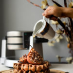 pouring maple syrup on a stack of brown waffles topped with baked pears and pecans with a waffle maker in the background