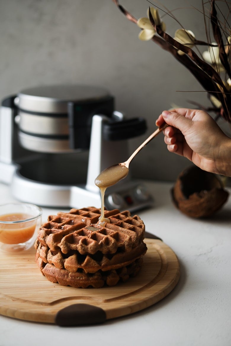 pouring caramel on a stack of brown waffles with a waffle maker in the background