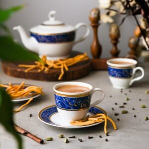 masala chai tea cup and saucer with a teapot in the background surrounded by plants and spices.