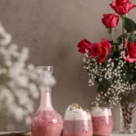 two glasses and a bottle of rose falooda in a tray with a backdrop of pink roses