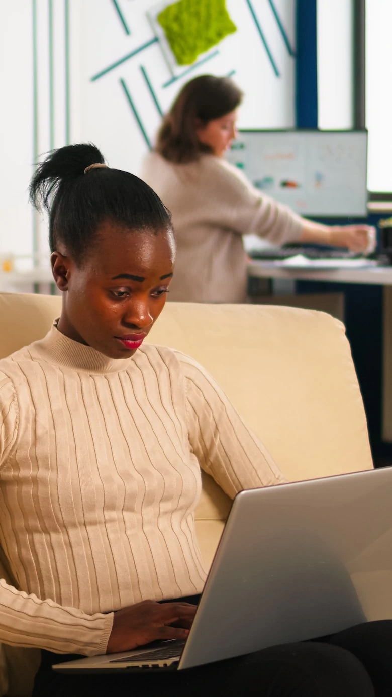 Concentrated african business woman answering emails typing on laptop sitting on couch in busy start up office while diverse team analyzes statistics data. Multiethnic team working at new project