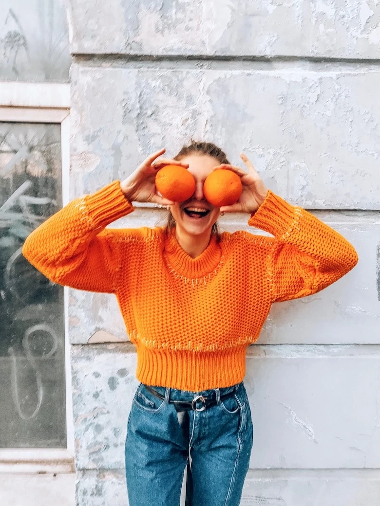 vertical, woman holding two oranges over her eyes while laughing, wearing a bright orange sweater