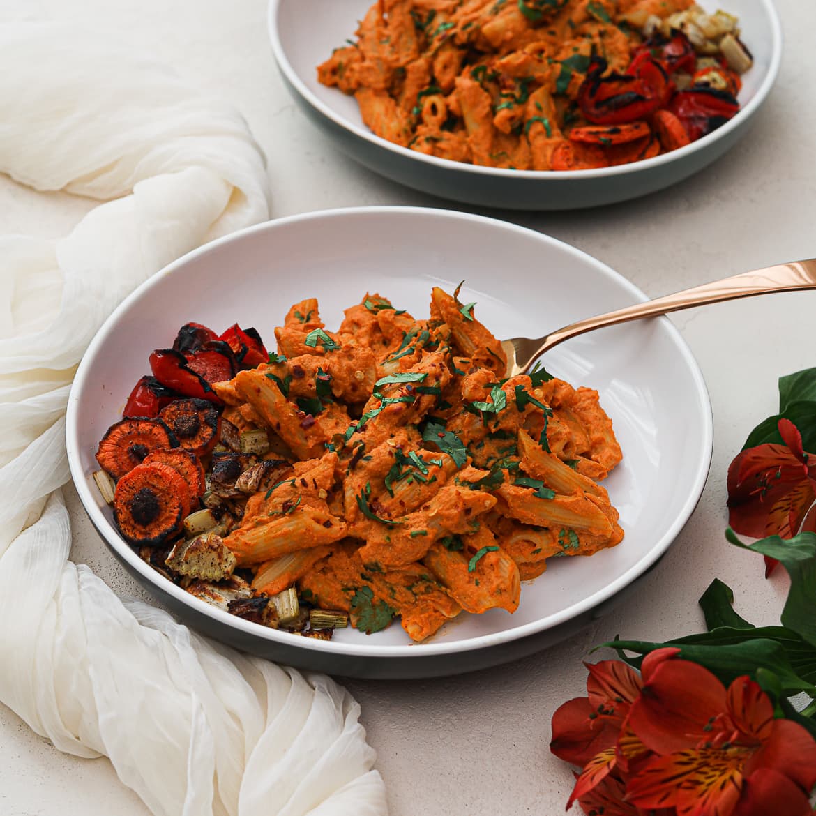 two bowls of creamy vegan pasta Indian style with a side of roasted vegetables and flowers in the foreground