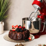 vegan chocolate cake bundt style on a stand topped with berries with a stand mixer in the background