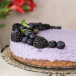 perspective shot of a blueberry dessert cake topped with black and blueberries, on a stand with flowers in the background.