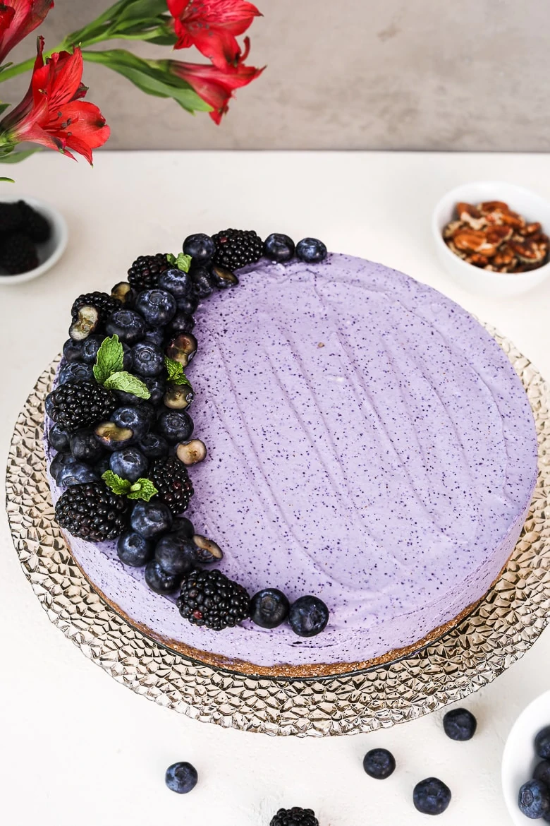 angle shot of a blueberry dessert cake topped with black and blueberries, on a stand with flowers and pecans in the background.