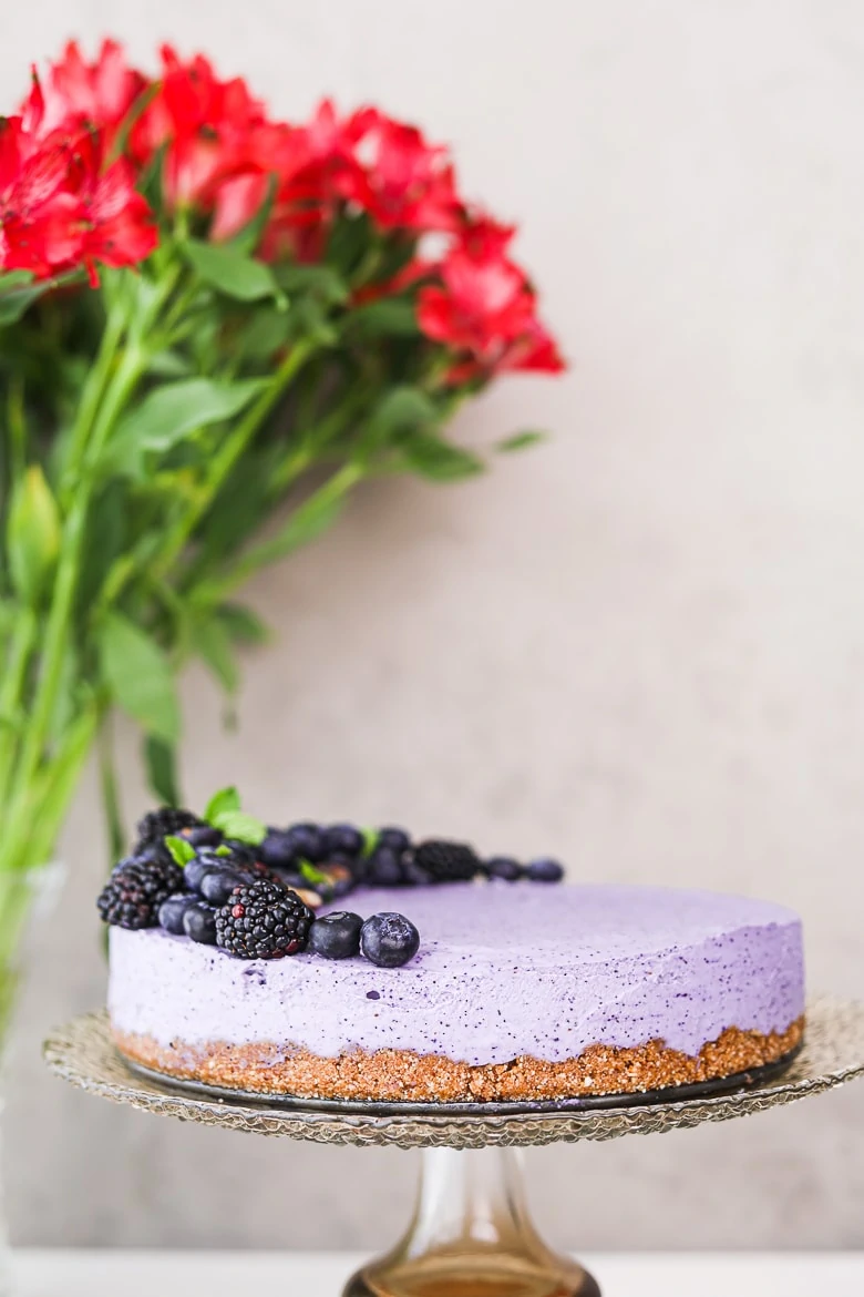 perspective shot of a blueberry dessert cake topped with black and blueberries, on a stand with flowers in the background.