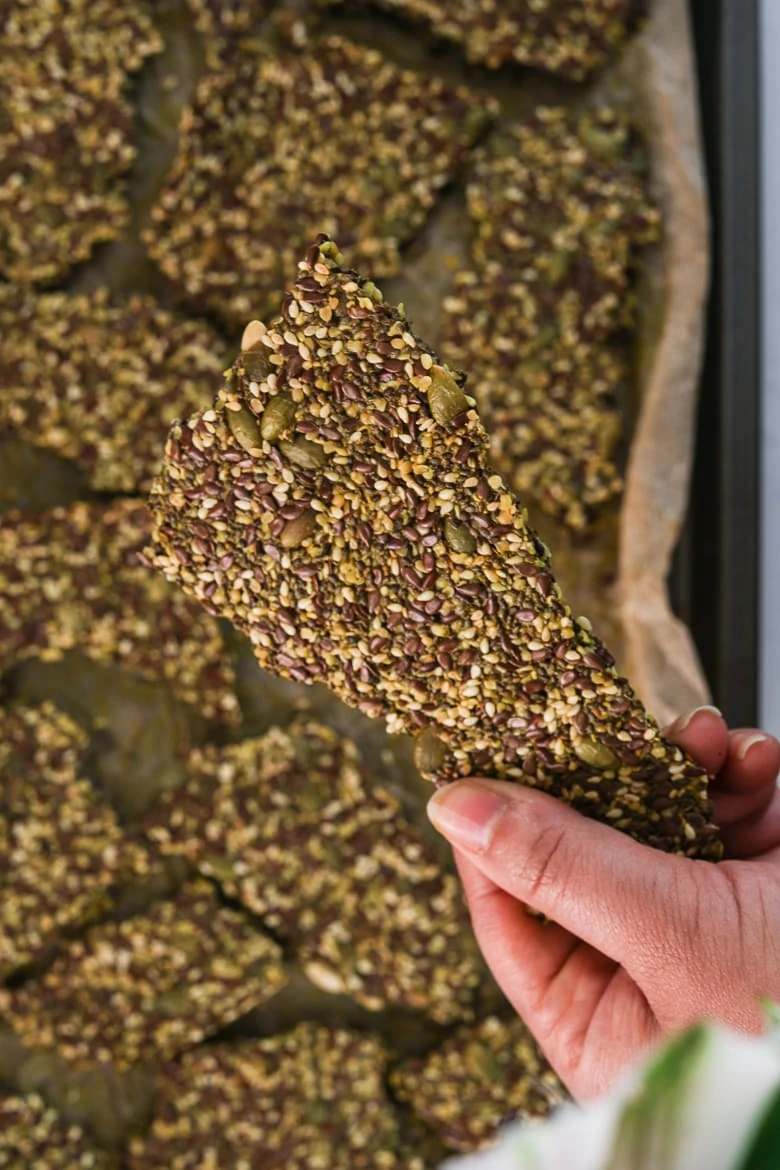 a hand holding a seeded cracker over a tray of backed seeded crackers of different shapes
