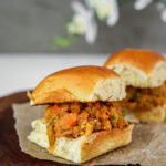 Indian pav bhaji filled in between two bread buns on a wooden board with white lily flowers in the background