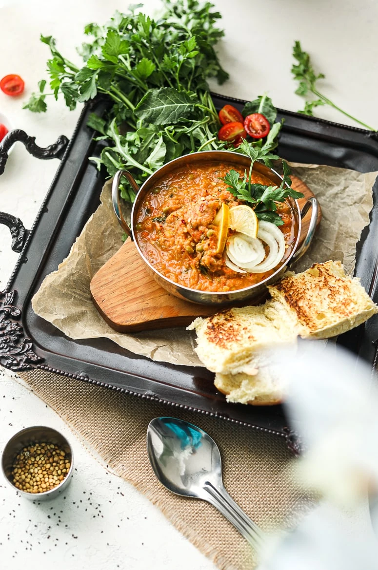 Bowl of Indian pav bhaji topped with onion slices and lemon segments on a black decorative tray with bread buns on the side and fresh herbs