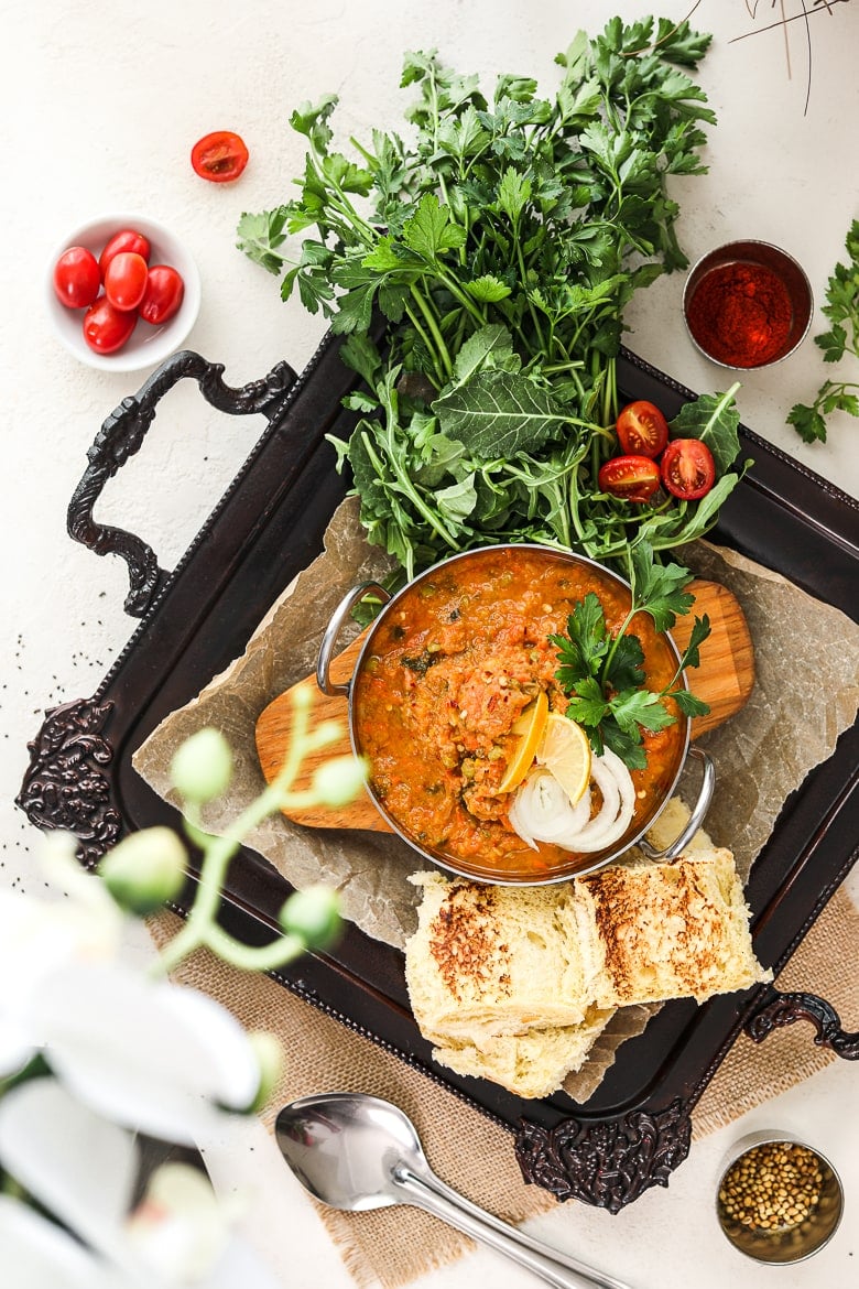 Bowl of Indian pav bhaji topped with onion slices and lemon segments on a black decorative tray with bread buns on the side and fresh herbs