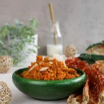 perspective shot of a green bowl with carrot halwa with a milk bottle in the background and wooden mesh balls in the foreground.