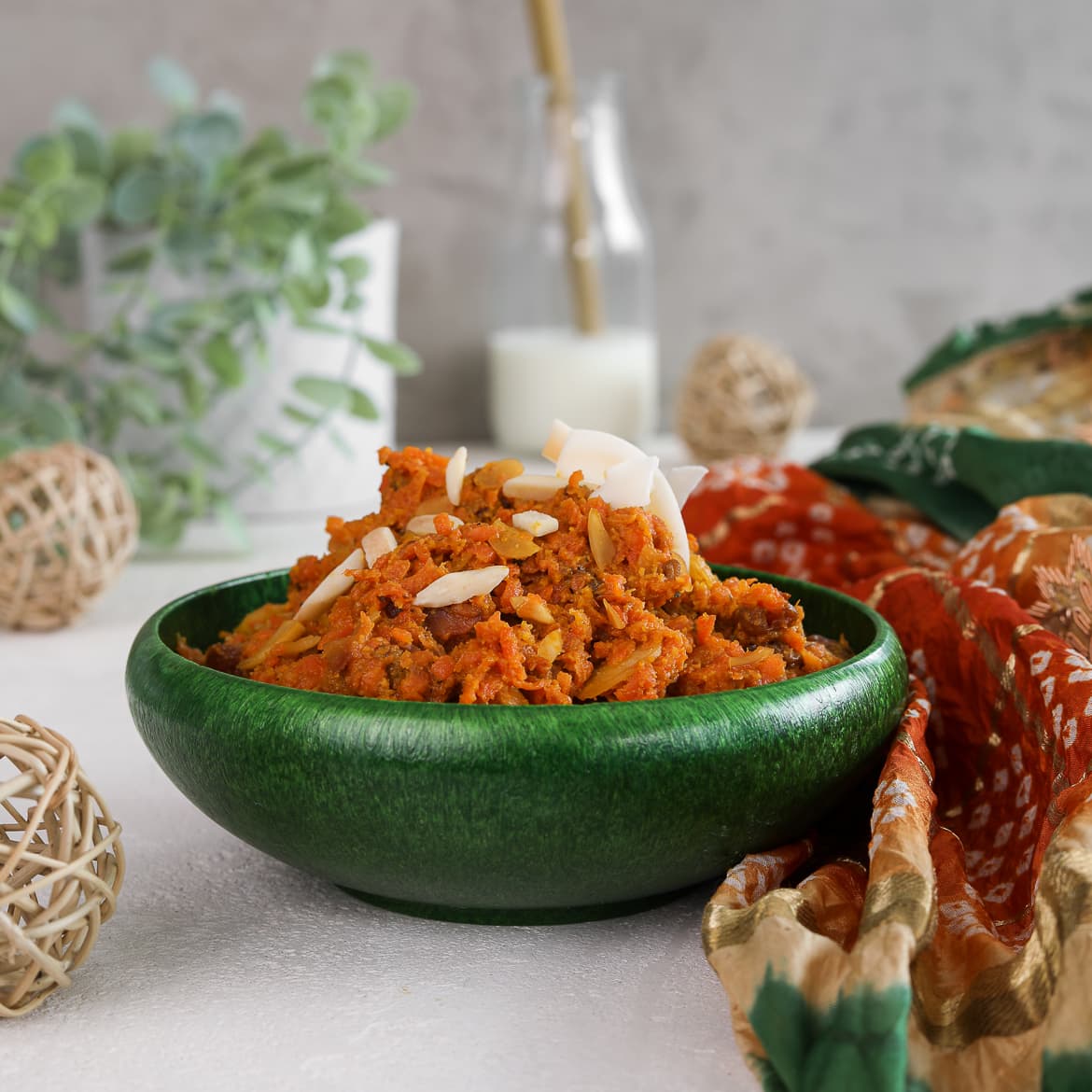perspective shot of a green bowl with carrot halwa with a milk bottle in the background and wooden mesh balls in the foreground with a traditional Indian shawl