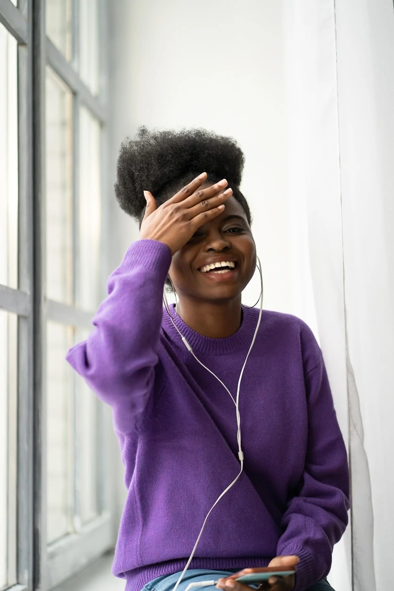African American millennial young woman with afro hairstyle wear purple sweater laughing with toothy smile, sitting on windowsill, listens to music with headphones on smartphone, looking at camera.