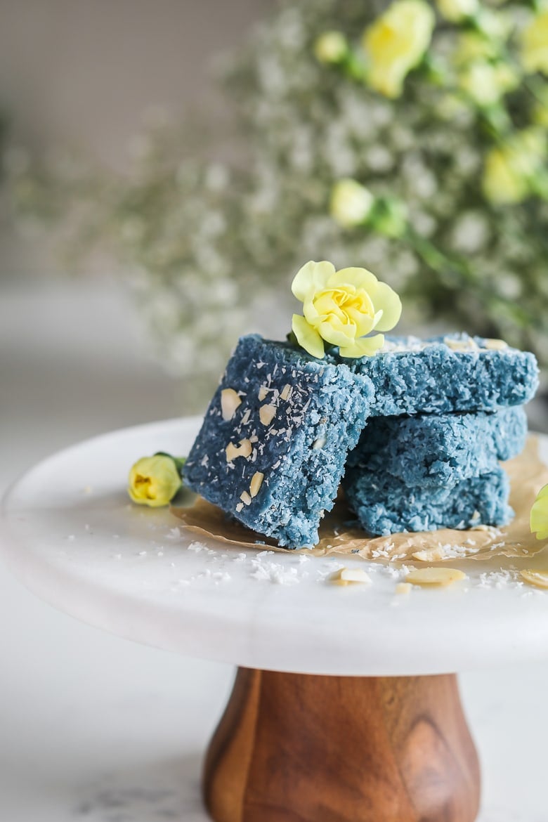 A stack of blue coconut barfi on a cake stand decorated with little yellow flowers
