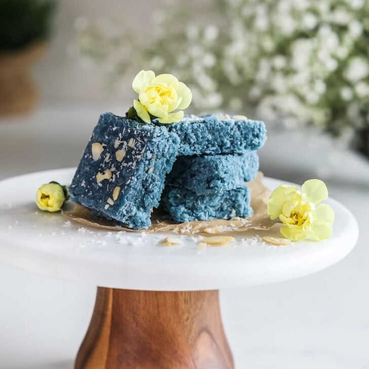 A stack of blue coconut barfi on a cake stand decorated with little yellow flowers