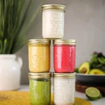 5 jars of colourful salad dressing recipes stacked on top of one another, on a yellow mat with a background of a bowl salad