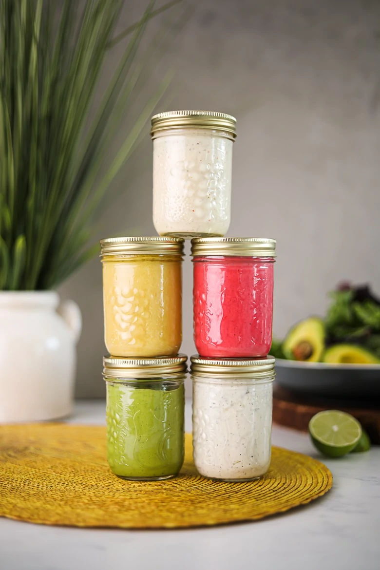 5 jars of colourful salad dressing recipes stacked on top of one another, on a yellow mat with a background of a bowl salad