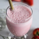 A glass of strawberry lassi with a bamboo straw with fresh strawberries in the background