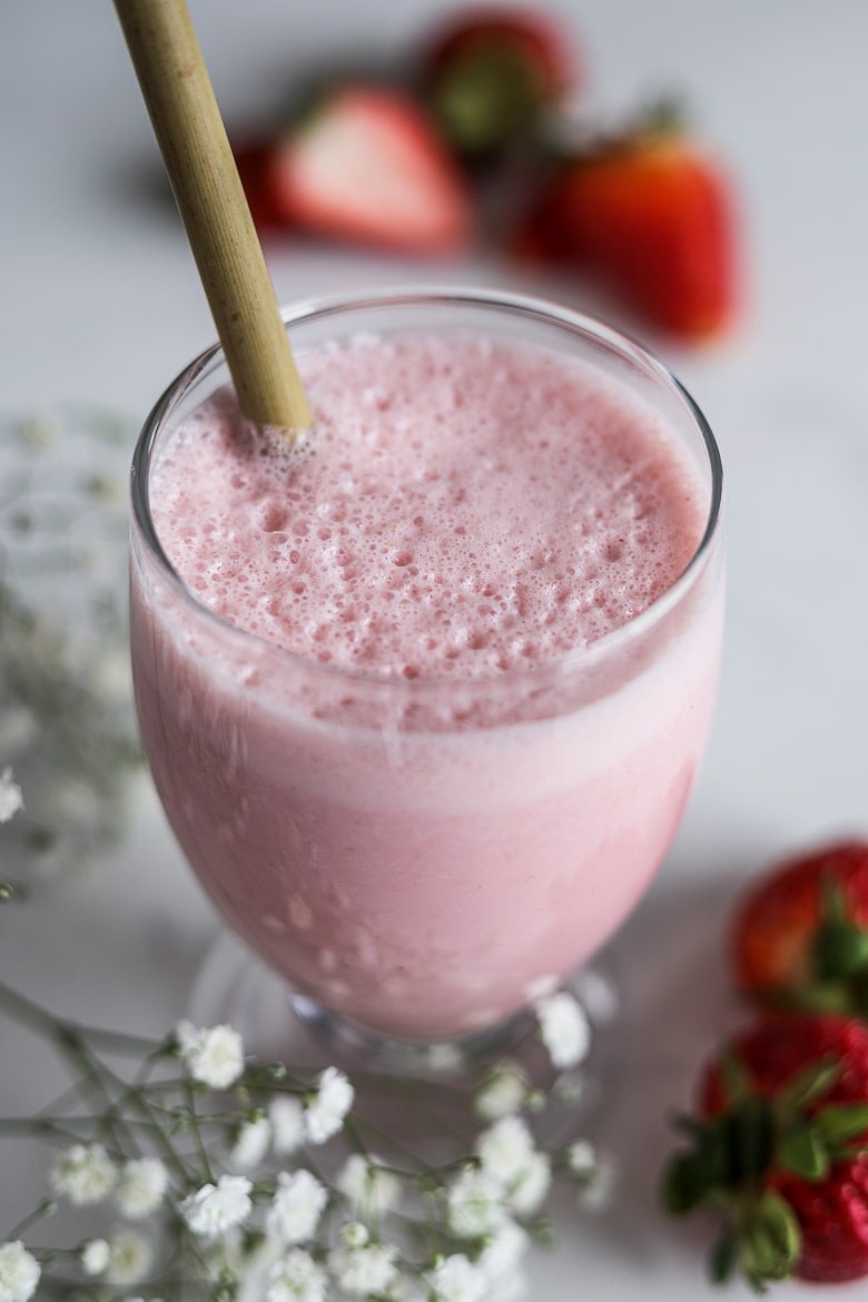 A glass of strawberry lassi with a bamboo straw with fresh strawberries in the background