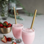 two glasses of strawberry lassi drink with bamboo straw dipped inside alongside a bowl of fresh strawberries with white flowers in the background