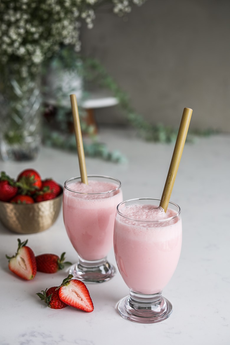 two glasses of strawberry lassi drink with bamboo straw dipped inside alongside a bowl of fresh strawberries with white flowers in the background